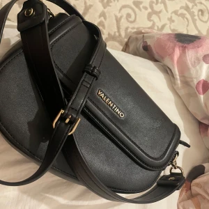 Svart axelväska från Valentino - Snygg svart axelväska från Valentino i skinn med justerbar axelrem och guldfärgade detaljer. Perfekt för att bära dina nödvändigheter med stil. Originala priset är 1300