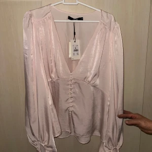 Ljusrosa blus med knappar - Elegant ljusrosa blus med djup v-ringning och knappar framtill. Blusen har långa ärmar med knytning vid ärmsluten och är tillverkad i ett glansigt material.