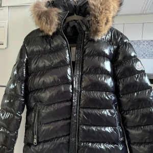 Svart dunjacka från Moncler - Snygg svart pufferjacka från Moncler med glansig yta och avtagbar huva med pälsdetalj. Jackan har en dragkedja framtill och två praktiska fickor med dragkedjor. Perfekt för kalla vinterdagar. Inte en enda skråma på jackan. Gammal modell så inget chip. Storlek 3/ passar mig som är M. Använd knappt en gång, då ja hittade en annan modell ja gillade mer :)