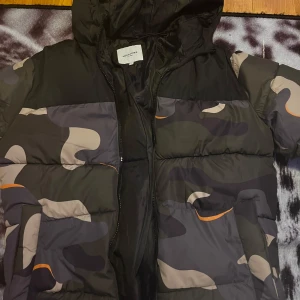 Camouflage pufferjacka från Jack & Jones - Säljer en cool pufferjacka från Jack & Jones i ett snyggt camouflage-mönster med inslag av orange. Jackan har en dragkedja framtill och en huva för extra skydd mot väder och vind.