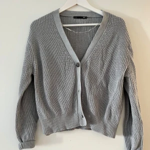 Grå stickad kofta från 157 - Säljer en mysig grå stickad kofta från 157. Den har en v-ringad design med knappar framtill och långa ärmar. Perfekt för kyliga dagar!