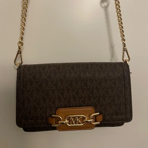 Brun axelväska från Michael Kors - Snygg brun axelväska från Michael Kors med guldkedja och logotypdetalj framtill. Väskan har flera kortfack och en dragkedja inuti, perfekt för att hålla ordning på dina saker. Den är stilren och passar till många olika tillfällen.