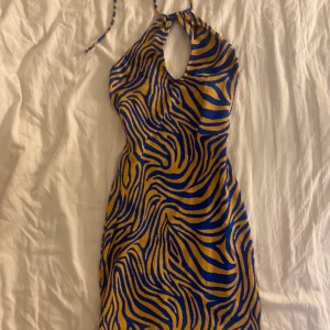 Zebramönstrad klänning med halterneck - Snygg zebramönstrad klänning i blått och gult med halterneck-design och öppen rygg. Klänningen har en unik öppning framtill och är perfekt för en djärv stil.