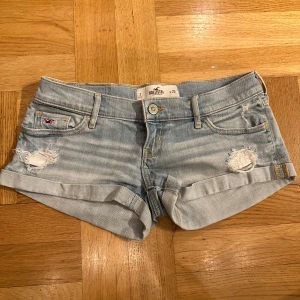 Ljusblå jeansshorts från Hollister - Snygga lågmidjade, ljusblå  jeansshorts med slitningar från Hollister. Klassisk femficksdesign och knappgylf. Midjemåttet är 40 cm!
