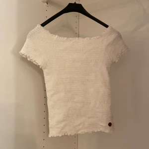 Vit ribbad topp från Ü - Säljer en söt vit ribbad topp från Ü i storlek 146/152. Toppen har korta ärmar och en elastisk offshoulder-design som ger en bekväm passform. Perfekt för varma sommardagar!