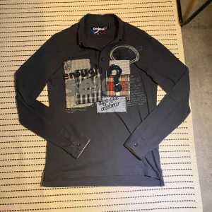 Svart långärmad pikétröja från Desigual - Säljer en svart långärmad pikétröja från Desigual med unika tryck och mönsterdetaljer i olika färger och stilar på framsidan. Tröjan har en klassisk krage och knappar framtill. Perfekt för dig som gillar en kreativ och annorlunda stil. Kan lösa bild med plagget på! (Pris kan diskuteras)