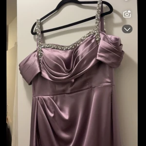 Ljuslila långklänning med glittriga axelband - Elegant ljuslila långklänning i satin med glittriga axelband och draperad överdel. Klänningen har en vacker slits framtill och offshoulder-design som ger en sofistikerad look. Original pris 5500 strl s/m