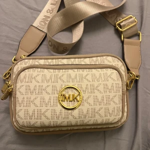 Beige axelväska  - Snygg beige axelväska  med logotypmönster och gulddetaljer. Väskan har en justerbar axelrem och flera dragkedjefack för praktisk förvaring. Perfekt för att bära med sig det viktigaste med stil.