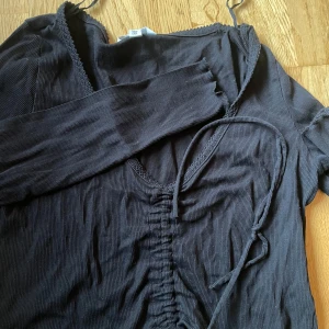 Svart ribbad topp med knyt - Säljer en svart ribbad topp med knyt framtill. Toppen har långa ärmar och en v-ringad design. Perfekt för en stilren look.