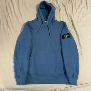 Blå hoodie från Stone Island - Säljer en snygg blå hoodie från Stone Island med deras ikoniska märke på ärmen. Tröjan har en klassisk känguruficka och justerbar huva med dragsko. Perfekt för en avslappnad stil.