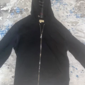 Burberry ziphoodie - Säljer en stilren svart huvtröja från Burberry med dragkedja och klassiskt rutigt mönster i fodret. Perfekt för en casual look med en touch av lyx. Huvan har ett snyggt mönster i grått och rött. Stringen för den finns men gick ba ut när jag hade tvättat den och att en av metallens snöre pä tröjan är sönder men det är ingenfara.