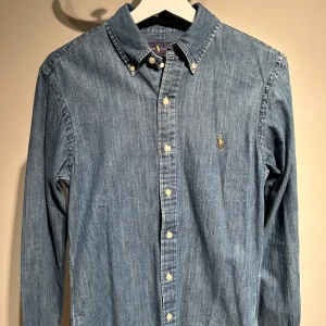 Blå jeansskjorta från Ralph Lauren - Snygg blå jeansskjorta från Ralph Lauren med klassisk krage och knappar framtill. Skjortan har en broderad logotyp på bröstet och är i slim fit. Perfekt för en stilren look.nypris 1000kr