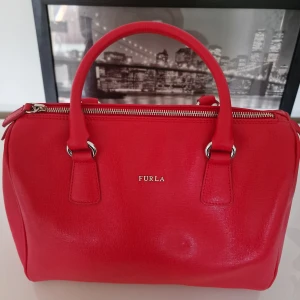 Röd handväska från Furla - Snygg röd handväska från italienska designmärket Furla med silverfärgade detaljer och dragkedja. Aldrig använd och därav säljes den. Garderobsrensning så kika gärna på övriga auktioner.