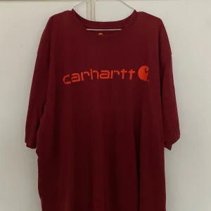 Vinröd t-shirt från Carhartt - Säljer en vinröd t-shirt från Carhartt med orange logotyp på framsidan. T-shirten har en normal passform och är kortärmad, perfekt för en avslappnad stil.