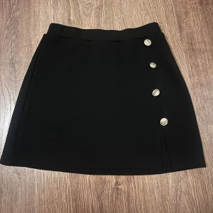 Svart kjol med dekorativa knappar - Elegant svart kortkjol med fyra dekorativa knappar i guld på sidan. Perfekt för en stilren look.