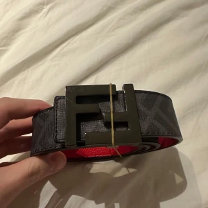 Svart och rött Fendi bälte med F-spänne - Snyggt bälte med ett stort F-spänne i svart. Bältet har en röd insida och ett diskret mönster på utsidan. Perfekt för att ge en stilren touch till din outfit.