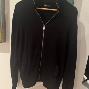 Svart kofta från Jack & Jones - Säljer en stilren svart kofta från Jack & Jones med dragkedja framtill. Perfekt för lager-på-lager under kyligare dagar. Enkel design som passar till det mesta.