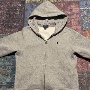 Grå hoodie från Ralph Lauren - Säljer en stilren grå hoodie från Ralph Lauren med dragkedja och huva. Tröjan har en liten broderad logga på bröstet och är perfekt för en avslappnad look. Den är i storlek S och passar både för kyliga dagar och mysiga stunder.