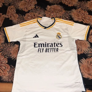 Vit Real Madrid fotbollströja - Säljer en vit Real Madrid fotbollströja från Adidas med gula och svarta detaljer. Tröjan har korta ärmar och klubbens emblem på bröstet. Perfekt för alla fans av laget!