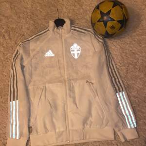 Säljer en snygg vit träningsjacka från Adidas med Sveriges landslagstryck. Jackan har dragkedja framtill och klassiska Adidas-ränder i reflekterande material längs ärmarna. Perfekt för fotbollsträning eller som en stilren vardagsjacka.