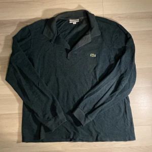 Mörkgrå pikétröja från Lacoste - Säljer en mörkgrön långärmad pikétröja från Lacoste. Tröjan har den ikoniska krokodilloggan på bröstet. Perfekt för en stilren och avslappnad look. Passar bra till både vardag och lite finare tillfällen.