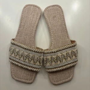 Beige sandaler med broderad detalj - Säljer ett par snygga beige sandaler med en fin broderad detalj på remmen. De har en fyrkantig tå och är perfekta för sommaren. Sandalerna är i syntetmaterial och har en bekväm passform.