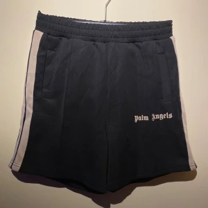 Svarta shorts från Palm Angels - Säljer ett par snygga svarta shorts från Palm Angels. De har en vit logga på framsidan och vita ränder längs sidorna. Shortsen har en elastisk midja och är perfekta för en avslappnad stil. De är helt nya och redo för sommaren!