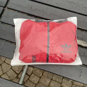 Säljer en snygg röd vindjacka från Adidas. Jackan har en dragkedja framtill och Adidas-loggan på bröstet, inklusive luva . Den har svarta detaljer och vita ränder längs ärmarna. Perfekt för vår och höst!    (NYPRIS ÄR 1899KR)