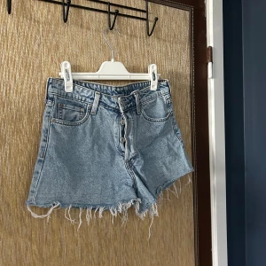 Blå jeansshorts med fransar - Säljer ett par snygga blå jeansshorts med fransiga kanter. De har en hög midja och knappar framtill. Perfekta för sommaren och ger en avslappnad look. Passar till både vardag och fest!