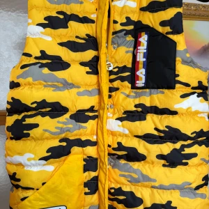 Gul kamouflagemönstrad dunväst från FILA - Säljer en supercool gul kamouflagemönstrad dunväst från FILA. Västen har en dragkedja framtill och en praktisk ficka med FILA-logga på bröstet. Perfekt för höst och vår när du vill sticka ut lite extra. Den är i nyskick och redo för nya äventyr!