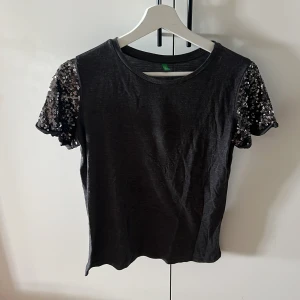 Svart topp med paljettärmar - Säljer en snygg mörkgrå topp med korta ärmar täckta av glittriga paljetter. Perfekt för festliga eller vardagliga tillfällen! Toppen är i ett mjukt och bekvämt material, och har en normal passform. Den är i bra skick och redo för nya äventyr.