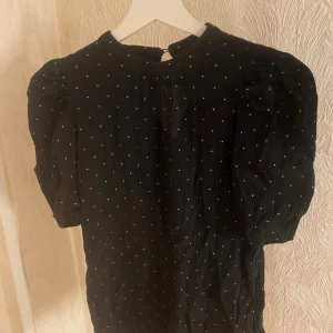 Svart blus med vita prickar - Säljer en söt svart blus med vita prickar. Blusen har korta puffärmar och en liten knappdetalj i nacken. Perfekt för både vardag och fest! 🖤