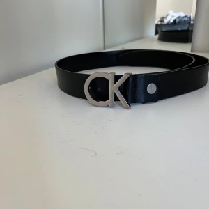 Svart bälte från Calvin Klein - Säljer ett stilrent svart bälte från Calvin Klein. Det har en snygg silverfärgad CK-logga som spänne. Bältet är 90cm långt och är av läder. Nypris 600.