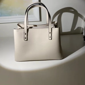 Beige handväska med dragkedja - Säljer en stilren beige handväska i syntetmaterial. Väskan har två handtag och en praktisk dragkedja upptill. Insidan är fodrad med ett prickigt mönster, vilket ger en fin kontrast. Perfekt för både vardag och fest!