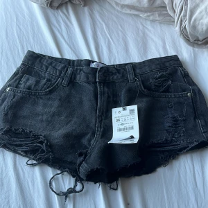 Svarta jeansshorts från Zara - Säljer ett par snygga svarta jeansshorts från Zara i storlek 36. De är lågmidjade och har en cool slitning på framsidan. Perfekta för sommaren och att matcha med en t-shirt eller linne. De är helt nya med prislappen kvar!