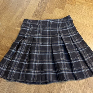 Brun rutig kjol med knappar - Säljer en superfin brun rutig kjol med knappar framtill. Kjolen har ett klassiskt rutmönster i brunt, beige och vitt. Perfekt för höst och vinter, och kan stylas både upp och ner beroende på tillfälle. Den har en normal passform och är i nyskick.