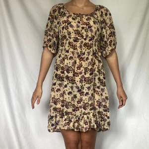 Blommig kortklänning med volangdetaljer - Säljer en superfin blommig kortklänning i beige med lila och bruna blommor. Klänningen har korta ärmar och en söt volangdetalj längst ner. Perfekt för sommarens alla fester eller en mysig dag på stan!