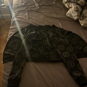 Svart tröja från Les Deux - Säljer en snygg svart tröj från Les Deux med ett subtilt mönster. Tröjan har dragkedja och långa ärmar, perfekt för en stilren look. Passar bra till både vardag och speciella tillfällen.