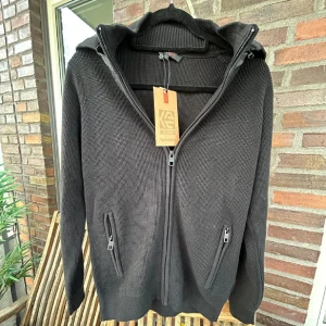 Svart stickad cardigan  - Snygg svart stickad cardigan med dragkedja och huva. Jackan har långa ärmar och praktiska fickor med dragkedjor. Perfekt nu när de kyligt ute och även den kommande våren.