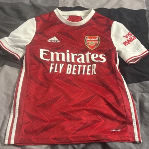 Röd Arsenal fotbollströja från Adidas - Säljer en röd Arsenal fotbollströja från Adidas med vitt mönster och korta ärmar. Tröjan har klubbens emblem på bröstet och sponsornamnet 'Emirates Fly Better' tryckt på framsidan. Perfekt för alla Arsenal-fans! ⚽️kontakta mig vid fler frågor😁😁😁passar 160-170cm 13-14 y 