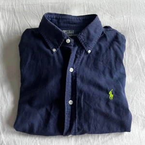 Mörkblå skjorta från Ralph Lauren - Säljer en stilren mörkblå skjorta från Ralph Lauren med klassisk krage och knappar framtill. Skjortan har en broderad grön logotyp på bröstet och är långärmad. Perfekt för en snygg och avslappnad look. Hör bara av dig vid någon fråga. Priset är inte hugget i sten