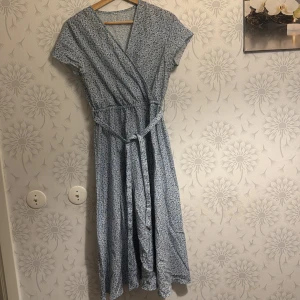 Blommig omlottklänning i blått - Charmig blå omlottklänning med blommigt mönster. Klänningen har korta ärmar och ett knytband i midjan för en smickrande passform. Perfekt för en somrig look!