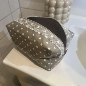 Grå prickig necessär med spetsdetalj - Charmig grå necessär med vita prickar och en söt spetsdetalj längs dragkedjan. Perfekt för att förvara smink eller andra småsaker. Praktisk och stilren design som passar i alla väskor💗💗