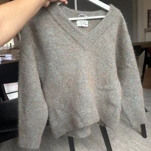 Beige tröja i ull och alpackablandning - Säljer en mysig  tröja i en mjuk ull- och alpackablandning. Färgen skiftar mellan grön-grå-brun.Tröjan har en v-ringad design och långa ärmar, perfekt för kyligare dagar. Den är i storlek L men känns mer som M har en avslappnad passform för den som har stl S. 