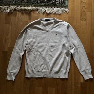 Beige tröja från Gant - Säljer en stilren beige tröja från Gant med v-ringning och broderad logga på bröstet. Tröjan har långa ärmar och ribbade kanter vid ärmslut och nederkant. Perfekt för en klassisk look.