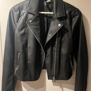 Svart skinnjacka med dragkedja - Snygg svart skinnjacka från H&M med coola dragkedjedetaljer och klassisk bikerstil. Jackan har en croppad passform och långa ärmar, perfekt för en edgy look. Passar utmärkt till både jeans och klänning. 🖤