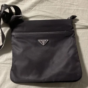 Svart axelväska från Prada - Snygg svart axelväska från Prada i syntetmaterial med justerbar axelrem. Väskan har en dragkedja upptill och ett elegant mönstrat foder inuti. Perfekt för att bära dina dagliga nödvändigheter med stil.