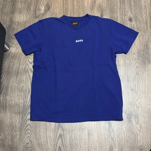 Blå t-shirt från Boss - Snygg blå t-shirt från Boss med korta ärmar och rund hals. Perfekt för en stilren och avslappnad look. Boss-loggan är diskret placerad på bröstet. Enkelt att matcha med jeans eller shorts.