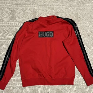 Röd tröja från Hugo - Snygg röd tröja från Hugo med svart logga på ryggen. Tröjan har svarta ränder med text längs ärmarna och en hög krage. Perfekt för en stilren look.