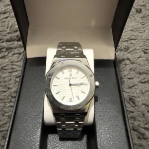 Silverfärgad klocka  - Elegant klocka i silver med en vit urtavla och stilrena indexmarkeringar. Armbandet är i rostfritt stål med AP-logotypen på spännet. Perfekt för den som söker en fin och lyxig accessoar.